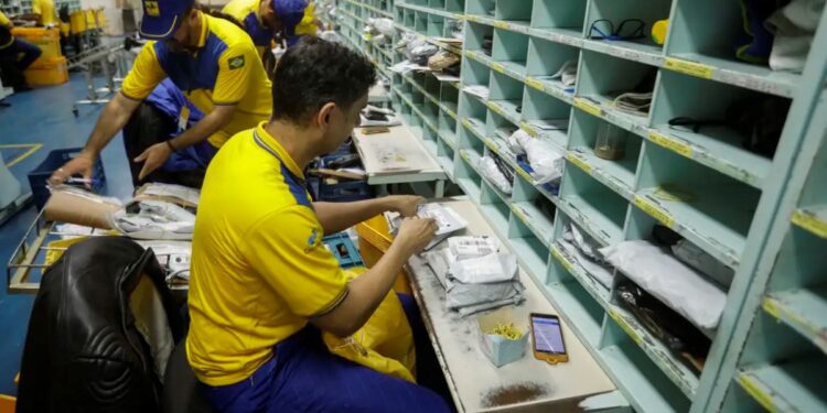 Funcionário dos Correios do Brasil manuseando e separando pacotes em uma bancada com caixas de correio ao fundo.