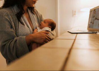 Mãe segurando recém-nascido enquanto visualiza informações sobre a ampliação do salário-maternidade em um laptop.