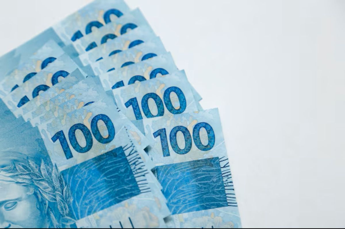 Várias cédulas de R$ 100,00 empilhadas em leque sobre uma superfície branca.