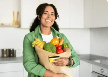 Pessoa sorrindo e segurando uma sacola de compras com frutas e legumes, representando a nova regulamentação do vale-alimentação em outubro.