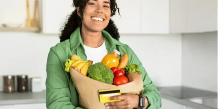 Pessoa sorrindo e segurando uma sacola de compras com frutas e legumes, representando a nova regulamentação do vale-alimentação em outubro.