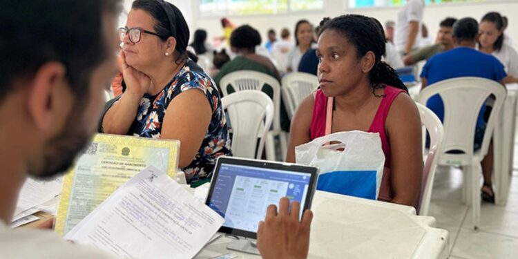 Imagem de mutirão de atendimento social para atualização do CadÚnico com pessoas sentadas em cadeiras plásticas, aguardando para serem atendidas. Em primeiro plano, uma mulher com expressão pensativa observa a tela de um tablet enquanto um técnico, com documentos em mãos, a auxilia.
