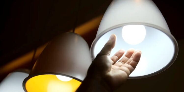 Mão tocando lâmpada acesa sob pendente branco, simbolizando o acesso à energia elétrica gratuita pelo programa Luz do Povo para famílias de baixa renda.