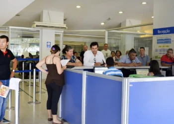 Pessoas em fila na recepção de um serviço público aguardando atendimento com funcionários em balcão azul