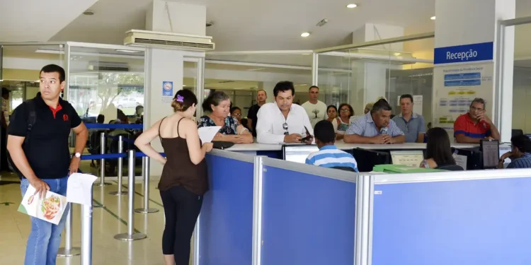 Pessoas em fila na recepção de um serviço público aguardando atendimento com funcionários em balcão azul