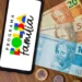 Celular mostrando logo do Programa Bolsa Família ao lado de notas e moedas de real sobre mesa de madeira