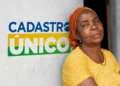 Mulher negra com lenço laranja e blusa amarela posa ao lado da frase Cadastro Único na parede