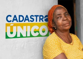 Mulher negra com lenço laranja e blusa amarela posa ao lado da frase Cadastro Único na parede