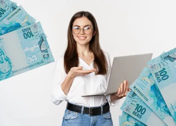 Mulher sorridente segura notebook, cercada por notas de 100 reais, representando datas de pagamento de benefícios como Bolsa Família e INSS em outubro.