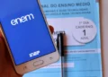 Celular com aplicativo do Enem aberto e caderno de prova azul do primeiro dia sobre mesa com caneta preta.