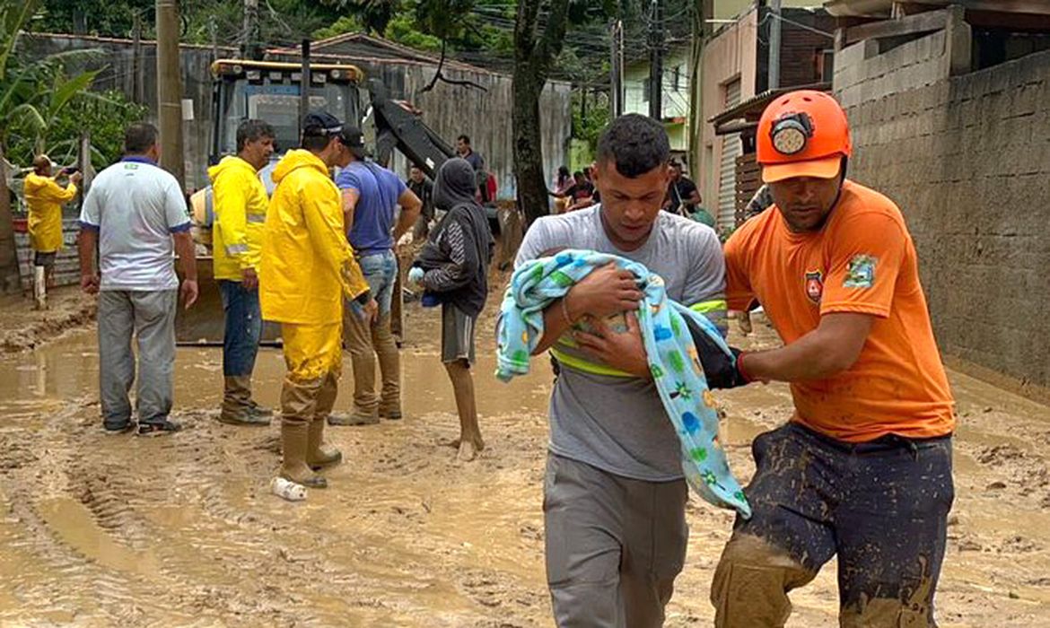 Homem carregando bebê enrolado em cobertor com várias pessoas em área alagada de lama e enchente