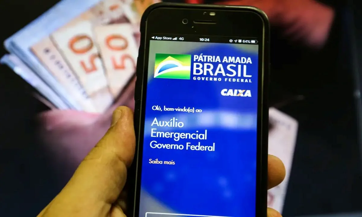 Tela do aplicativo Caixa Econômica mostrando a mensagem de Auxílio Emergencial, enquanto dinheiro em cédulas é visível ao fundo.
