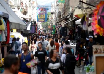 Multidão de pessoas caminhando em uma rua movimentada de feira ou mercado ao ar livre, com barracas e produtos, refletindo a atmosfera de feriados e lazer.