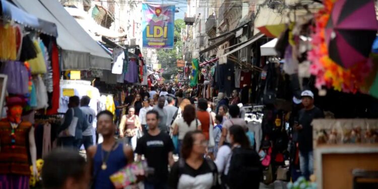 Multidão de pessoas caminhando em uma rua movimentada de feira ou mercado ao ar livre, com barracas e produtos, refletindo a atmosfera de feriados e lazer.