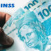 Mão segurando notas de 100 reais com o logo do INSS ao fundo.