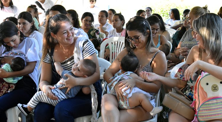 Mulheres sentadas em cadeiras brancas amamentando seus bebês em espaço externo durante evento