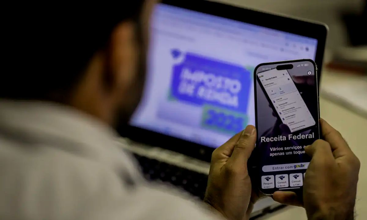 Consulta Restituição IR 2025 Pessoa consultando a restituição do Imposto de Renda em um aplicativo da Receita Federal no smartphone, com um notebook ao fundo.