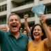 Grupo de pessoas sorrindo e comemorando enquanto uma mulher segura várias cédulas de dinheiro azul