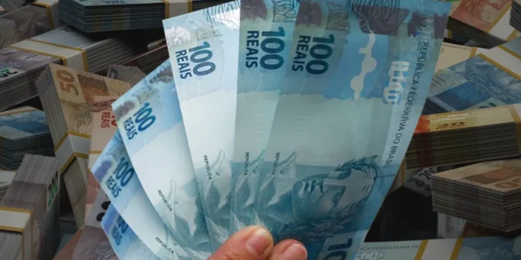 Mão segurando um leque de notas de R$ 100, com pilhas de dinheiro em segundo plano, representando um abono ou saque de valores.