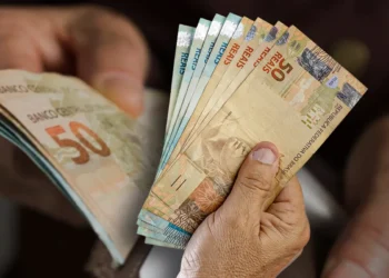 Pessoa contando várias cédulas de dinheiro em reais na mão, com cédulas de 50 e 20 reais visíveis