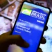 Smartphone exibindo app oficial do Auxílio Emergencial do Governo Federal em fundo de cédulas de reais