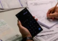 Pessoa usando calculadora no celular e escrevendo valores em caderno para controlar contas e despesas