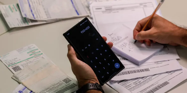 Pessoa usando calculadora no celular e escrevendo valores em caderno para controlar contas e despesas