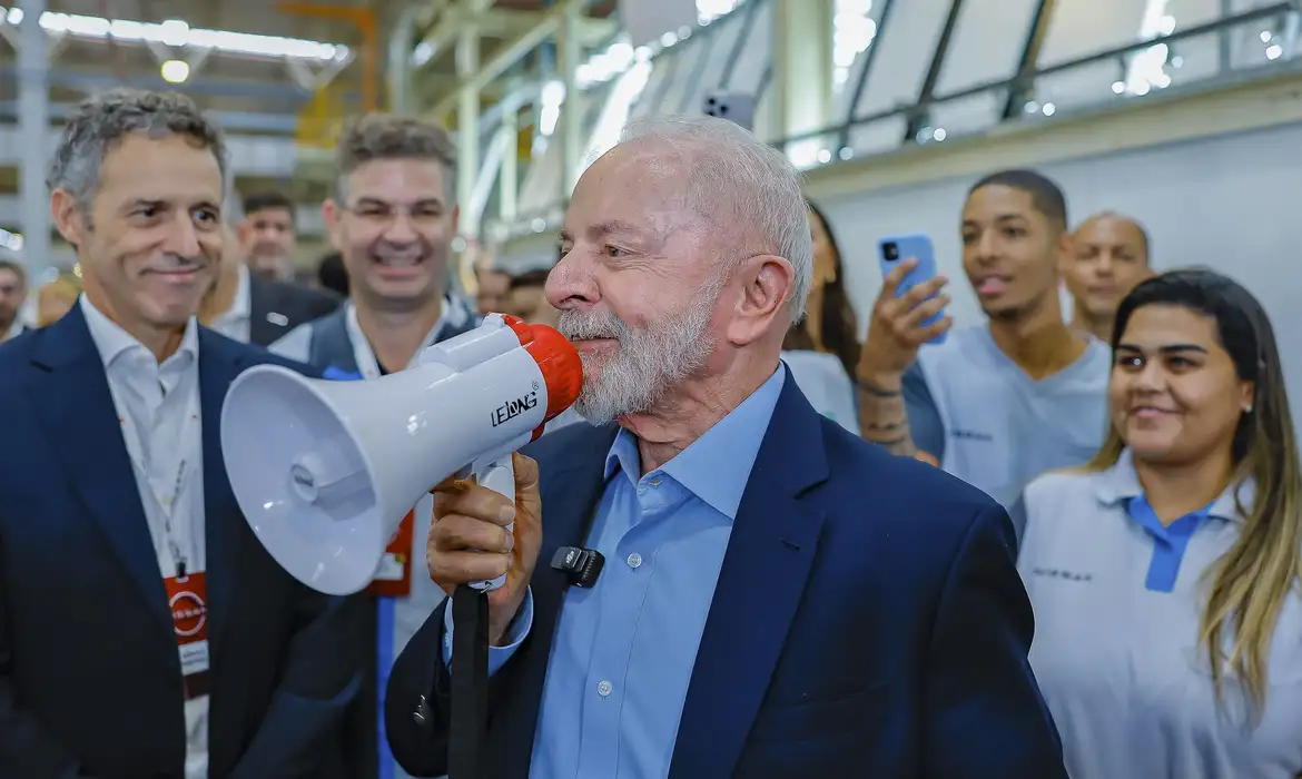 Político falando em megafone durante evento público Homem usando megafone fala com pessoas ao redor em evento público dentro de espaço com iluminação natural