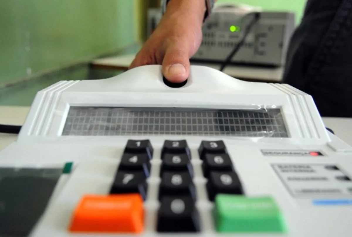Mão apertando botão biométrico na urna eletrônica branca com teclas coloridas em escritório.