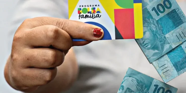 Cartão do Bolsa Família na mão de pessoa com notas de cem reais ao fundo.
