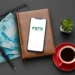 Imagem de um celular com o logo do FGTS sobre um fundo de dinheiro, caneta, planta e café. Representa a consulta ao FGTS de forma profissional e moderna.