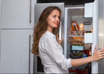 Mulher de camisa branca sorrindo ao abrir a porta de uma geladeira na cozinha moderna