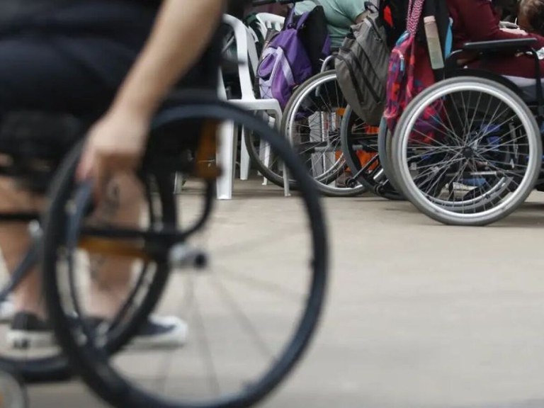 Pessoas em cadeiras de rodas em ambiente interno. Várias pessoas utilizando cadeiras de rodas em ambiente interno com mochilas e cadeira plástica ao fundo.