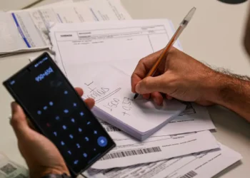 Pessoa calculando contas com caneta e papel e usando calculadora no celular