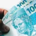Mão segurando notas de 100 reais, moeda brasileira, em 2026.