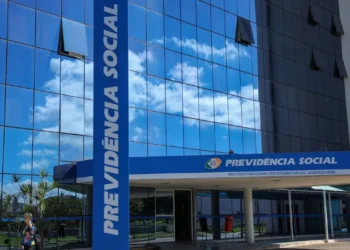 Fachada do edifício sede do INSS, com a placa 'Previdência Social' visível e um céu azul refletido nas janelas.
