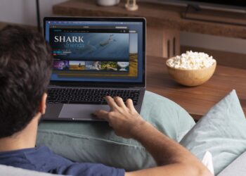 Homem sentado no sofá mexendo no notebook assistindo documentário Shark Friends com tigela de pipoca ao lado