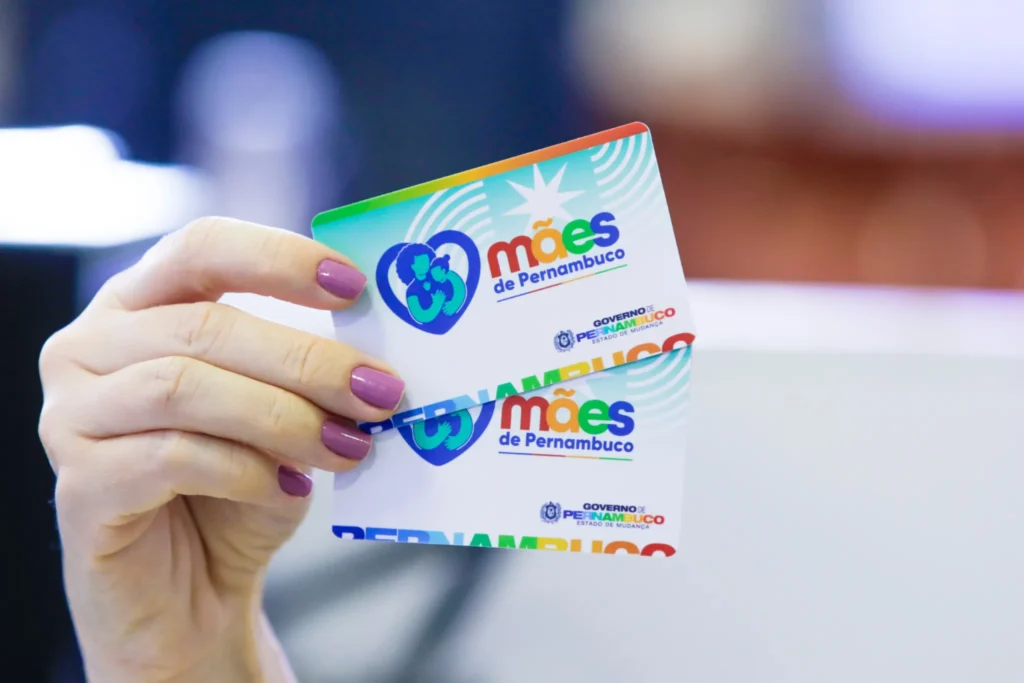 Cartões do programa “Mães de Pernambuco” em destaque. Mão segurando dois cartões do programa “Mães de Pernambuco”.