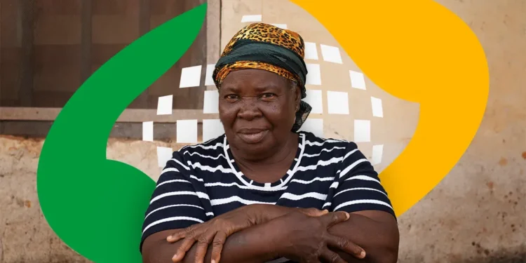 Mulher idosa com blusa listrada e lenço na cabeça, posando com os braços cruzados em frente a um fundo de parede com formas abstratas.