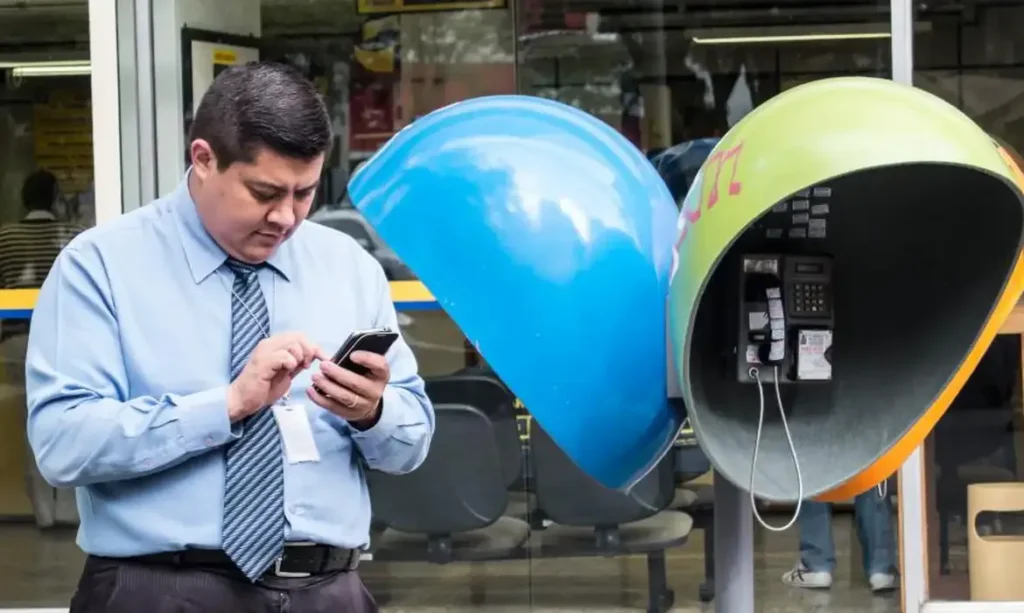 O fim dos orelhões e a evolução da telefonia. Homem utilizando um smartphone ao lado de um orelhão colorido, representando a transição da telefonia pública para a mobilidade no Brasil.