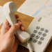 Mão segurando telefone fixo branco sobre mesa com mapas no fundo