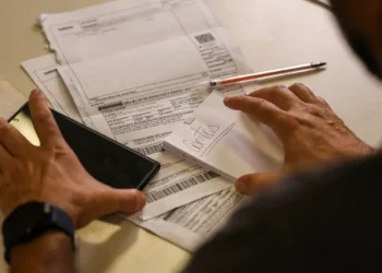 Mãos manuseando celular, boletos, bloco anotado como contas e caneta sobre mesa