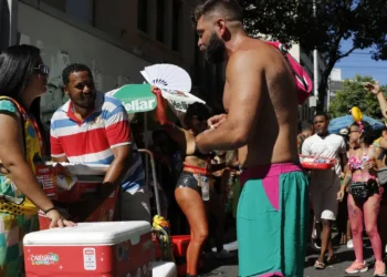 Homens e mulheres vendem bebidas geladas para foliões fantasiados em rua ensolarada