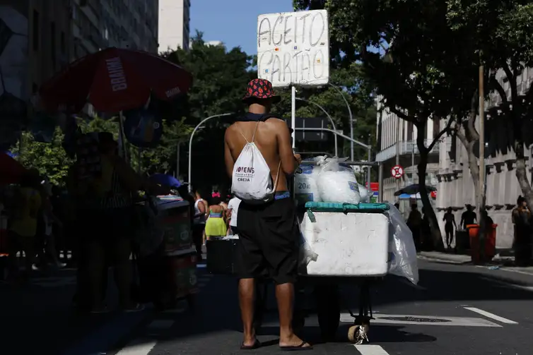 Ambulante vendendo gelo durante Carnaval de rua em 2026 Homem empurra carrinho de gelo com placa “Aceito cartão” em rua movimentada