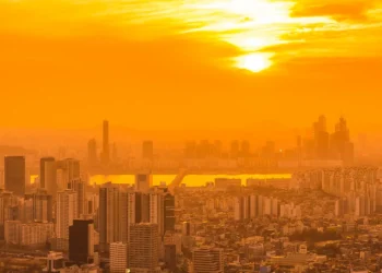 Prédios altos de cidade grande banhados por intensa luz alaranjada do pôr do sol em dia de calor extremo.