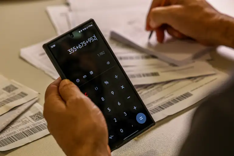 Pessoa usando celular para calcular contas no início de 2026 Mão segura celular com calculadora aberta enquanto outra faz anotações perto de boletos