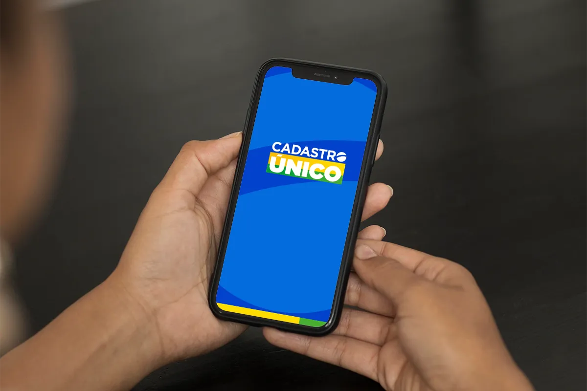 Tela do Cadastro Único exibida em smartphone para atualização cadastral 2026. Pessoa segura celular com tela azul do aplicativo Cadastro Único destacada para atualização de dados.