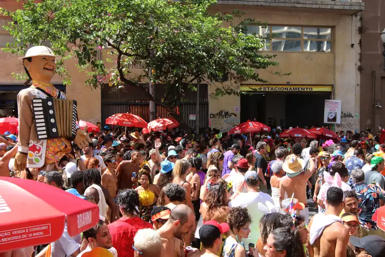 Boneco de Olinda e foliões animam bloco de rua no Carnaval 2026 Multidão de foliões fantasiados festeja sob sol, boneco de Olinda e guarda-sóis vermelhos presentes
