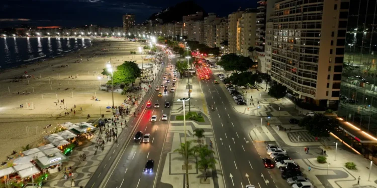 Carros trafegando em avenida à noite junto à praia iluminada e prédios residenciais.