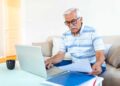 Idoso sentado no sofá usando notebook enquanto confere documentos